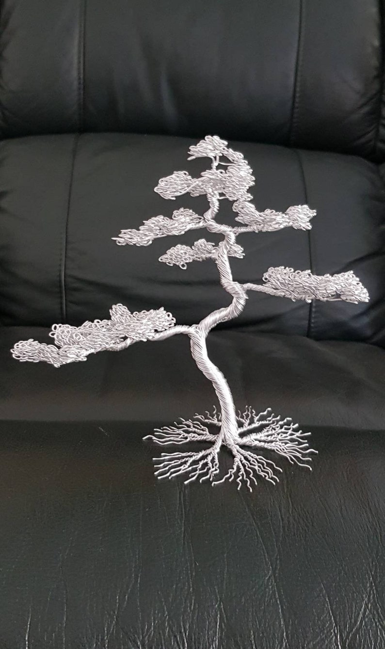 May include: A silver wire sculpture of a bonsai tree. The tree has a twisted trunk, branches with textured foliage, and exposed roots. The sculpture is displayed on a black surface.