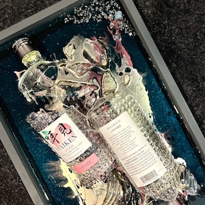 May include: Two clear glass bottles of Haiken Lychee liqueur, encased in a teal blue resin with gold and pink swirls. The bottles are partially submerged in the resin, with the labels facing up. The labels read "Haiken Lychee" and "Lychee Crafted in Japan".