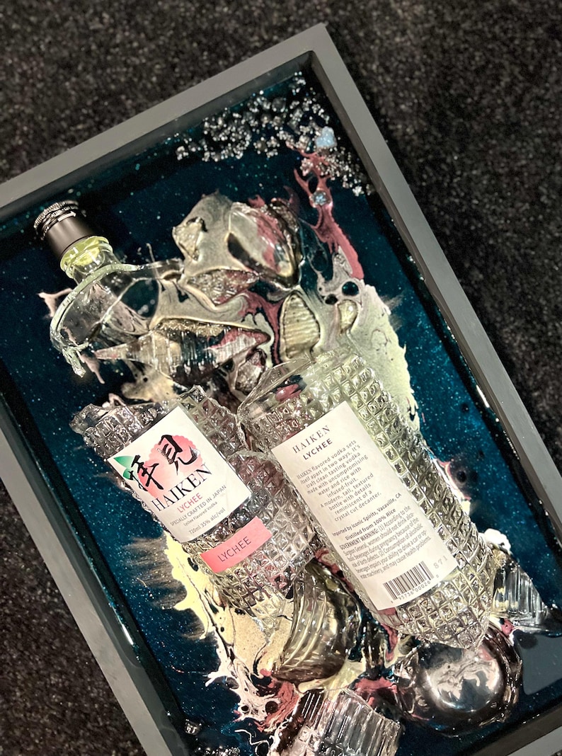 May include: Two clear glass bottles of Haiken Lychee liqueur, encased in a teal blue resin with gold and pink swirls. The bottles are partially submerged in the resin, with the labels facing up. The labels read "Haiken Lychee" and "Lychee Crafted in Japan".