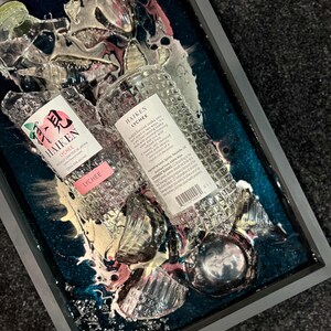 May include: Two clear glass bottles of Haiken Lychee flavored vodka, partially submerged in a teal blue resin with pink and white swirls. The bottles are broken and shattered, with the labels facing up. The text on the labels reads "Haiken Lychee" and "Finely Crafted in Japan."