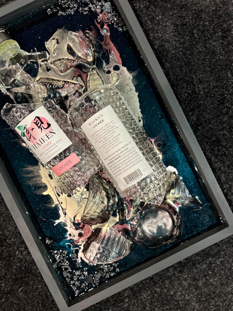 May include: Two clear glass bottles of Haiken Lychee flavored vodka, partially submerged in a teal blue resin with pink and white swirls. The bottles are broken and shattered, with the labels facing up. The text on the labels reads "Haiken Lychee" and "Finely Crafted in Japan."