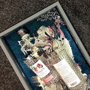 May include: Two clear glass bottles of Haiken Lychee liqueur, one upright and one laying down, are encased in a teal blue resin with gold and pink swirls. The bottles are partially submerged in the resin, with the labels visible. The resin is set in a gray frame.
