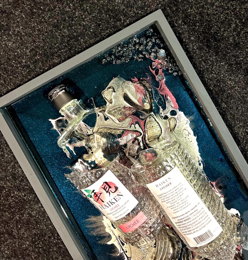 May include: Two clear glass bottles of Haiken Lychee liqueur, one upright and one laying down, are encased in a teal blue resin with gold and pink swirls. The bottles are partially submerged in the resin, with the labels visible. The resin is set in a gray frame.