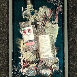 May include: A framed artwork featuring two clear glass bottles of Haiken Lychee liqueur, partially submerged in a teal blue resin with silver and pink swirls. The bottles are surrounded by broken glass shards and have a textured, diamond-like pattern.