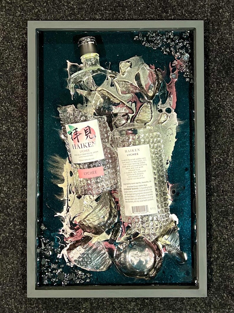 May include: A framed artwork featuring two clear glass bottles of Haiken Lychee liqueur, partially submerged in a teal blue resin with silver and pink swirls. The bottles are surrounded by broken glass shards and have a textured, diamond-like pattern.