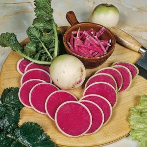 May include: A white radish with a pink interior is sliced into thin rounds. The radish is surrounded by green leaves and a small bowl of shredded pink radish.
