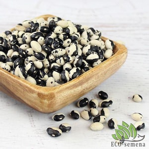 以下が含まれることがあります： 黒と白の斑点のある豆が木製のボウルに入っています。豆は白い木製の表面に散らばっています。