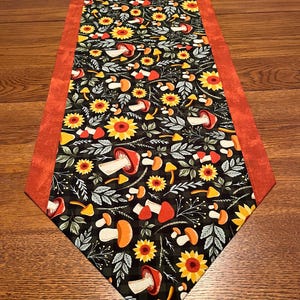 May include: A decorative table runner featuring a black background with a pattern of sunflowers, mushrooms, and leaves in shades of orange, yellow, and white. The runner has a solid orange border and is placed on a wooden table.