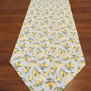May include: A rectangular table runner with a repeating pattern of yellow lemons, green leaves, and small blue flowers on a light gray background. The runner is made of fabric and is placed on a wooden table.