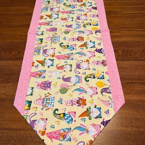 May include: A decorative Easter-themed table runner. The center features a yellow fabric with colorful gnome and bunny illustrations. Pink patterned fabric borders the runner's edges. The runner is designed to be placed on a table.
