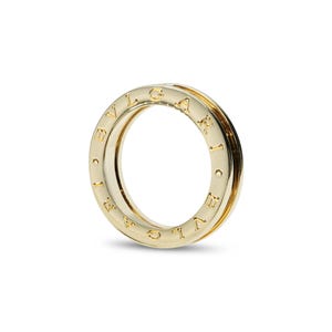 May include: A polished gold ring featuring a thick, cylindrical band with a hollow center. The outer surface of the ring is engraved with the text BVLGARI in capital letters, repeating around the circumference of the metallic gold band.