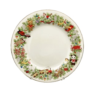 May include: A white ceramic plate with a gold rim features a festive Christmas scene with toys, a rocking horse, a teddy bear, a bicycle, and a panda bear. The plate is decorated with green holly leaves and red berries.