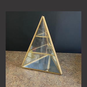 May include: A geometric terrarium in a pyramid shape. The structure is made of clear glass panels and a gold-colored metal frame. The terrarium is designed with interior shelves for displaying small plants or decorative items. It sits on a brown surface.