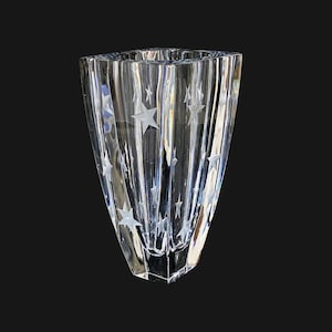 May include: A clear, faceted glass vase with a star pattern. The vase has a flared shape and vertical ridges. The stars are etched into the glass, creating a subtle, elegant design. The vase is suitable for floral arrangements or as a decorative piece.