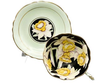 May include: A vintage teacup and saucer set with a black background and yellow daffodils painted on it. The saucer is a pale green color with a gold rim.