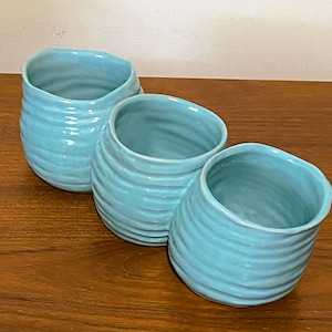 May include: Three light blue ceramic planters with a textured, ridged design. The planters are arranged in a row, each with a unique oval shape. The glossy finish reflects light, highlighting the curves and the aqua color.