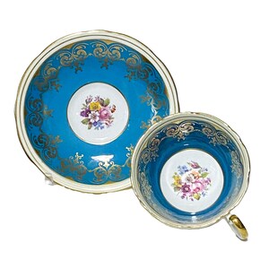 May include: A vintage teacup and saucer set. The set features a vibrant turquoise blue background with gold filigree detailing. The center of the cup and saucer has a white circle with a floral bouquet. The cup has a gold handle.