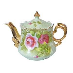 May include: A vintage porcelain teapot with a light green base, adorned with pink and red roses and gold accents. The teapot features a curved spout, a looped handle, and a lid with a gold finial. The floral design is hand-painted.