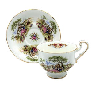 May include: A white teacup and saucer set with a gold rim. The cup and saucer feature a pastoral scene with figures in pink, blue, and green. The set is likely made of porcelain.