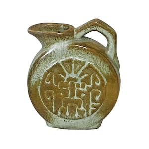 May include: A small, round ceramic pitcher with a handle and spout. The pitcher is primarily brown with a lighter, textured glaze around the top and a decorative emblem on the front. The emblem is a sunburst design with geometric shapes.