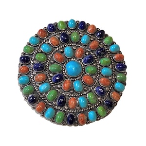May include: A round, silver-toned brooch with a central turquoise stone surrounded by concentric circles of colorful stones. The stones include turquoise, orange, green, and dark blue, creating a vibrant, eye-catching design. The brooch has a detailed, textured border.