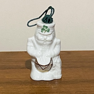 May include: A white ceramic figurine of a leprechaun drummer, with a green shamrock on his hat. The figurine has a gold-trimmed drum and a green cord for hanging. A festive Irish-themed decoration.