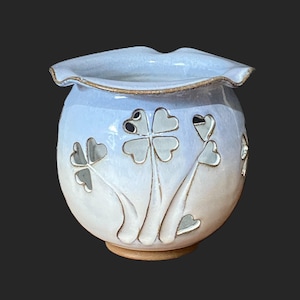 May include: A ceramic, round vase with a light blue and white gradient. The vase features four-leaf clover cutouts with gold-colored outlines. The rim has a ruffled edge and a gold-colored trim. The base is a light brown color.