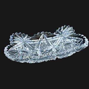 May include: A clear, oval-shaped crystal dish featuring intricate cut-glass patterns. The surface is adorned with geometric starburst motifs and cross-hatched detailing, with a scalloped, serrated edge that catches the light across its entire body.