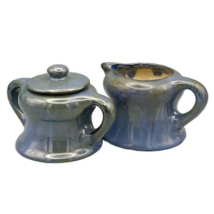 May include: A blue and silver ceramic sugar bowl with a lid and a matching creamer. The set has a glossy finish and a unique, rounded shape. The creamer has a handle and a spout.