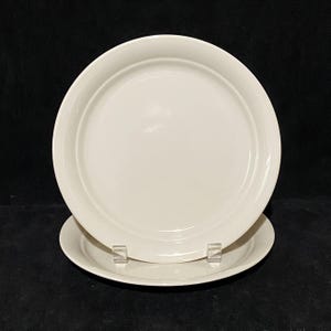 May include: Two off-white ceramic plates are stacked on a black surface. The top plate is slightly angled, revealing the rim and the flat center. The bottom plate is partially visible, with a similar design. The plates are simple and elegant.