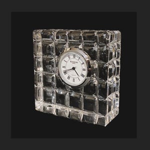 May include: A clear, rectangular crystal clock with a square-cut pattern. The clock face is white with black Roman numerals and hands, and the words "Waterford Crystal Quartz" are printed on the face. The clock has a silver-colored bezel.