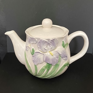 May include: A white ceramic teapot with a lid and handle. The teapot is decorated with raised relief iris flowers in shades of purple, green, and yellow. The teapot has a spout and a round knob on the lid.