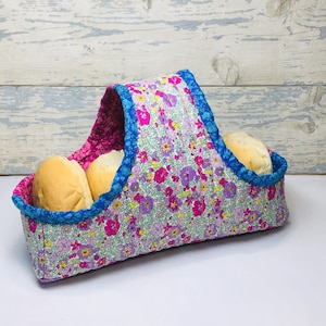 Peut inclure: Un panier à pain en tissu floral rose et bleu avec une poignée. Le panier est rempli de trois pains ronds.