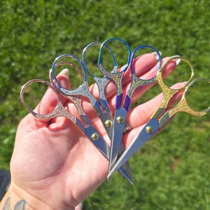 May include: A set of four small metal scissors with ornate, textured handles in varying metallic finishes including rose gold, silver, iridescent blue-green, and gold. Each pair features a central brass screw and sharp, pointed steel blades.