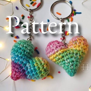 Puede incluir: Dos llaveros de coraz&oacute;n de crochet arco&iacute;ris con anillas de plata. Los corazones est&aacute;n hechos con una variedad de colores, incluyendo rosa, azul, verde y amarillo. La palabra "Pattern" est&aacute; escrita en blanco sobre un fondo blanco.