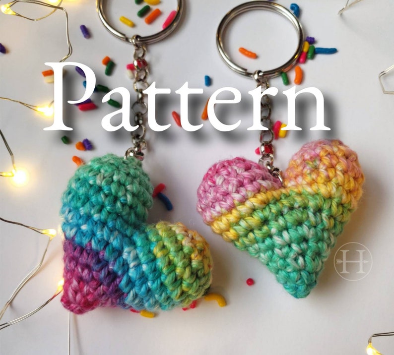 Puede incluir: Dos llaveros de coraz&oacute;n de crochet arco&iacute;ris con anillas de plata. Los corazones est&aacute;n hechos con una variedad de colores, incluyendo rosa, azul, verde y amarillo. La palabra "Pattern" est&aacute; escrita en blanco sobre un fondo blanco.
