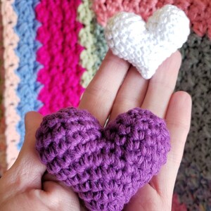 Puede incluir: Dos corazones tejidos a crochet, uno morado y otro blanco, se sostienen en una mano. El texto "Swooning Hearts Pattern makes the most perfect little puffy hearts." est&aacute; debajo de los corazones.