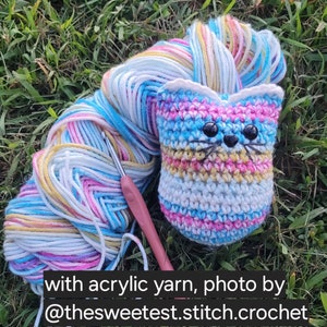 May include: A crocheted cat toy with a striped pattern in pink, blue, white, and yellow. The cat toy has black eyes and a black nose. A ball of multi-colored yarn and a crochet hook are also in the image. with acrylic yarn, photo by @thesweetest.stitch.crochet