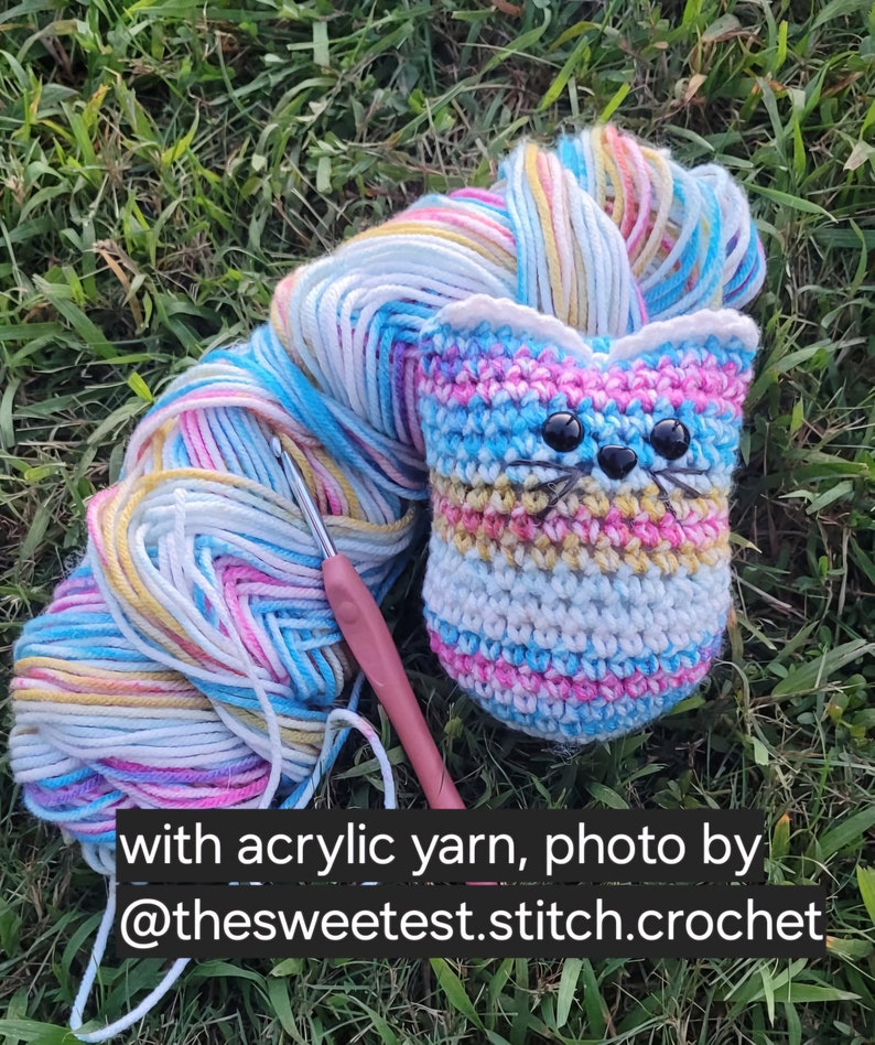 May include: A crocheted cat toy with a striped pattern in pink, blue, white, and yellow. The cat toy has black eyes and a black nose. A ball of multi-colored yarn and a crochet hook are also in the image. with acrylic yarn, photo by @thesweetest.stitch.crochet