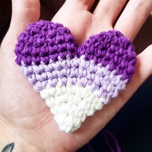 Puede incluir: Un coraz&oacute;n de crochet en tonos de morado y blanco. El coraz&oacute;n est&aacute; hecho con un punto texturizado y tiene una forma ligeramente irregular.