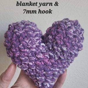 Puede incluir: Una almohada de punto de crochet en forma de coraz&oacute;n hecha con hilo morado, rosa y blanco. La almohada est&aacute; hecha con un gancho de crochet de 7 mm. El texto "blanket yarn & 7mm hook" est&aacute; escrito encima de la almohada.