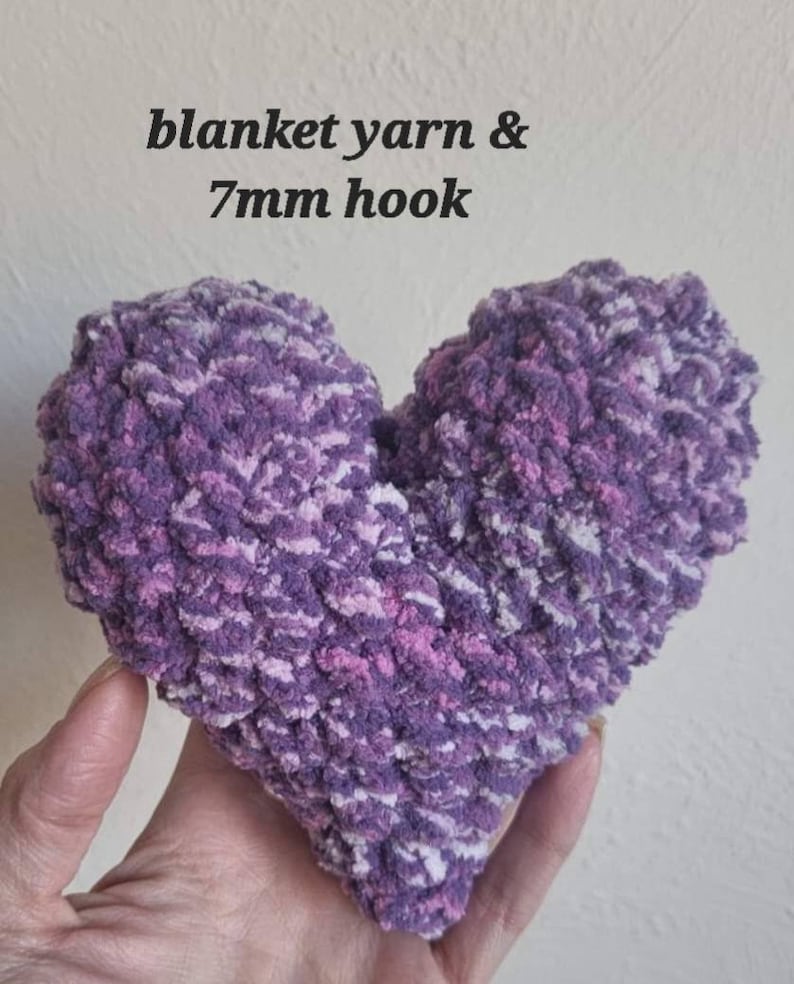 Puede incluir: Una almohada de punto de crochet en forma de coraz&oacute;n hecha con hilo morado, rosa y blanco. La almohada est&aacute; hecha con un gancho de crochet de 7 mm. El texto "blanket yarn & 7mm hook" est&aacute; escrito encima de la almohada.