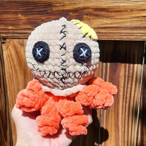 May include: A crocheted plush toy with a stitched face, orange limbs, and a yellow patch on its head. The toy is being held in a hand with a butterfly tattoo.