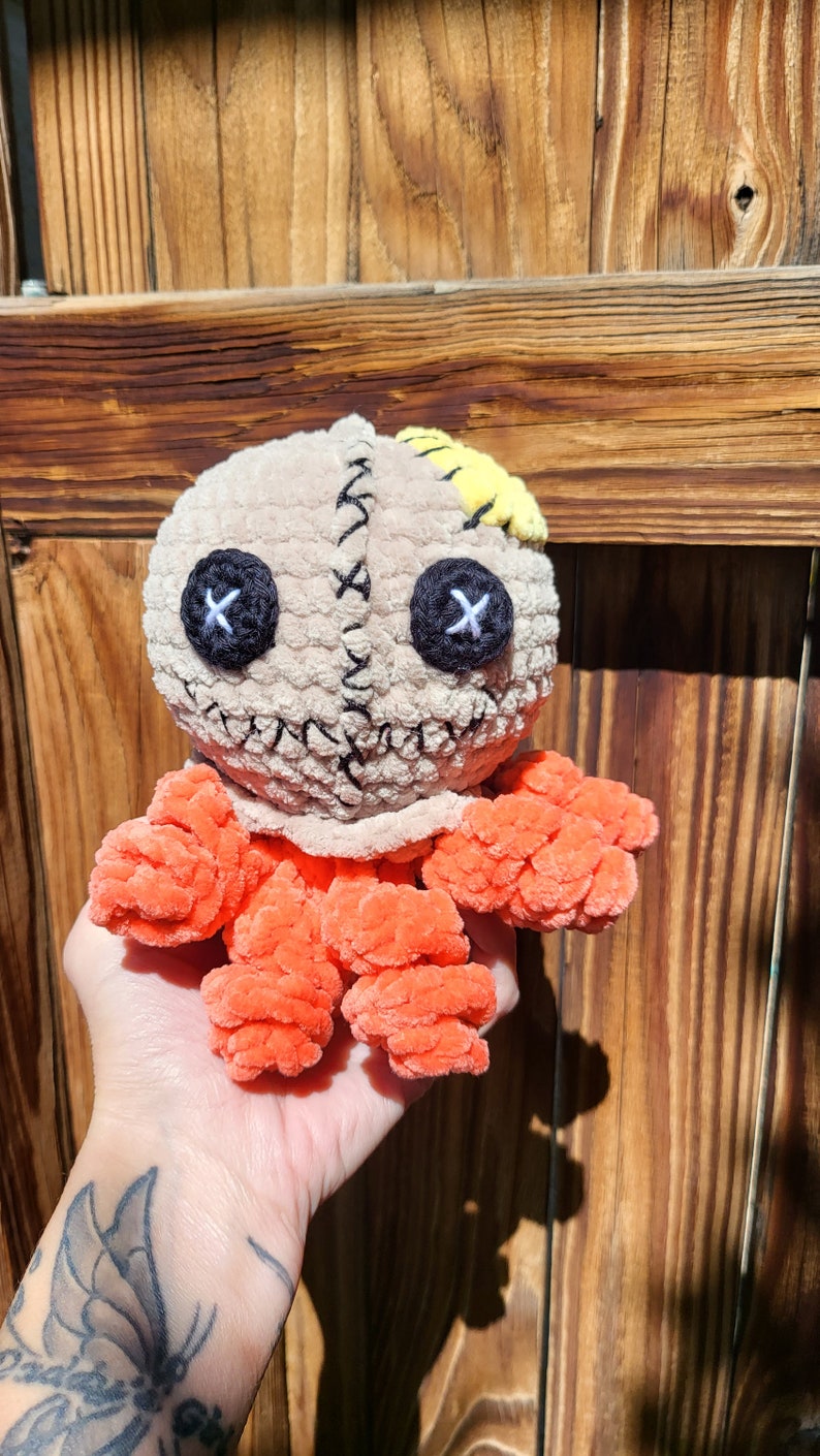 May include: A crocheted plush toy with a stitched face, orange limbs, and a yellow patch on its head. The toy is being held in a hand with a butterfly tattoo.