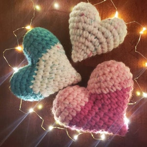 Puede incluir: Tres corazones de ganchillo en diferentes combinaciones de colores: azul verdoso y blanco, rosa y rojo, y blanco con motas multicolores. Los corazones est&aacute;n dispuestos en c&iacute;rculo sobre una superficie marr&oacute;n con una cadena de luces blancas alrededor.