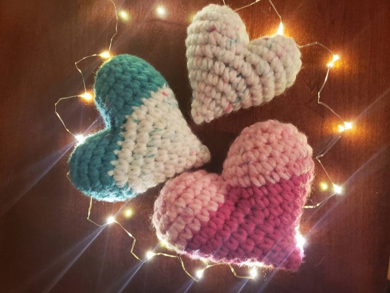 Puede incluir: Tres corazones de ganchillo en diferentes combinaciones de colores: azul verdoso y blanco, rosa y rojo, y blanco con motas multicolores. Los corazones est&aacute;n dispuestos en c&iacute;rculo sobre una superficie marr&oacute;n con una cadena de luces blancas alrededor.
