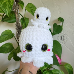 May include: White crocheted octopus plush toy with large green eyes and pink cheeks. A smaller white crocheted octopus with green eyes sits on top of the larger octopus.