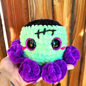 May include: Green and black crocheted octopus with purple tentacles. The octopus has a stitched-on Frankenstein face with black eyes and pink cheeks.