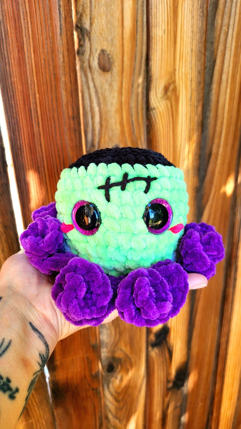 May include: Green and black crocheted octopus with purple tentacles. The octopus has a stitched-on Frankenstein face with black eyes and pink cheeks.