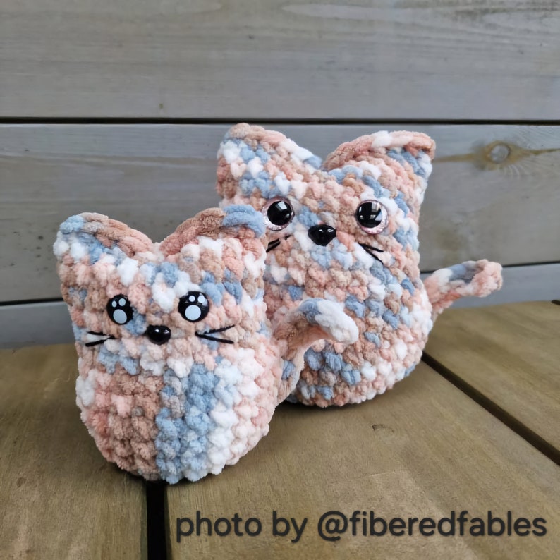 May include: Two crocheted cat plushies in a light blue, pink, and white color scheme. The plushies have black eyes and whiskers.